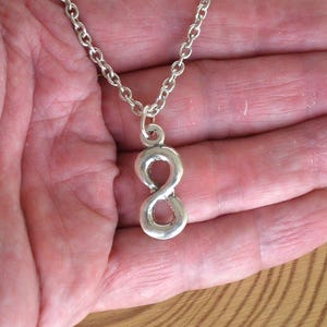 Puede incluir: Collar con colgante de símbolo de infinito plateado. El colgante, un diseño de figura de ocho estilizada, cuelga de una delicada cadena. El collar se muestra sobre un fondo neutro, resaltando el diseño y el brillo metálico de la joya.