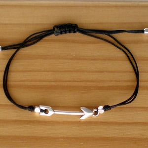 Può includere: Un braccialetto in cordoncino nero con un ciondolo a forma di freccia d'argento. Il braccialetto è regolabile e ha un design semplice.