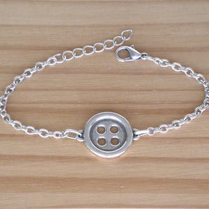 Puede incluir: Una pulsera plateada con un dije en forma de botón con cuatro agujeros. La pulsera tiene un diseño de eslabones de cadena y una longitud ajustable con un cierre de mosquetón. La joya se exhibe sobre una superficie de madera marrón claro.