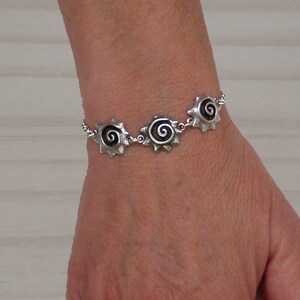 Op de afbeelding: Een zilveren armband met drie bedels met een spiraalvormig ontwerp. De bedels zijn verbonden door een zilveren ketting.