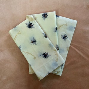 May include: Three beige reusable food wraps with a bee and floral design and the text "Bumble" printed on them.