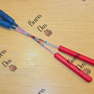 May include: Two pairs of juggling clubs are displayed on a light wood surface. One pair has blue handles and the other has red handles. The clubs have silver shafts with colorful accents.
