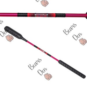 Könnte beinhalten: Eine rosa-schwarze Reitpeitsche mit einem schwarzen Ledergriff und einem rot-schwarz-weißen gemusterten Schaft. Die Peitsche ist mit "Bears Den" in grauer Schrift beschriftet.