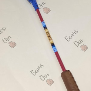 May include: A long, slender rod featuring a blue leather-wrapped handle at the top and a brown leather-wrapped handle at the bottom. The central red shaft is accented with a decorative gold-toned metal segment and horizontal stripes in light blue.