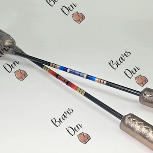 Könnte beinhalten: Zwei schwarze Steigbügel mit dekorativen Griffen. Die Steigbügel haben farbenfrohe Perlenakzente in Rot, Gold und Blau. Der Text "Bears Den" wiederholt sich über das Bild.