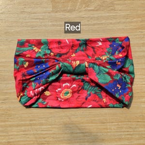 Può includere: Una fascia larga in tessuto con un vivace motivo floreale composto da grandi fiori rossi, fiori blu e foglie verdi. Il tessuto è arricciato al centro per creare un effetto a nodo. La parola Red è stampata in bianco su uno sfondo grigio scuro.
