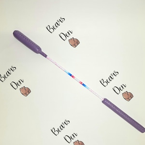 Könnte beinhalten: Ein lila-weißer Zauberstab mit einem farbenfrohen Streifendesign. Der Zauberstab hat abgerundete Griffe und einen verzierten Schaft. Die Worte "Bears Den" sind in schwarzer Schrift um den Zauberstab geschrieben.