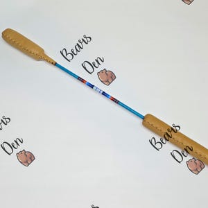 May include: A tan leather and beaded stick with a blue shaft. The stick has tan leather ends and a central section adorned with red, white, and blue beads. The words "Bears Den" are printed on the background.