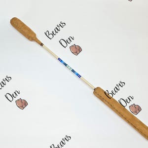 Könnte beinhalten: Ein hellbraunes Lederpaddel mit einem hellen Holzgriff und blauen und weißen Perlen. Das Paddel hat eine abgerundete Schlagfläche und einen rechteckigen Griff. Die Worte "Bears Den" sind im Hintergrund geschrieben.