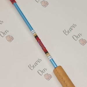 May include: A long, slender rod featuring a bright blue metallic shaft, a red textured handle at the top, and a tan leather-wrapped grip at the bottom. The center of the shaft displays a decorative section with red, black, and gold bands.