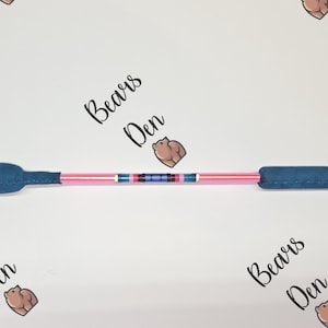 Könnte beinhalten: Eine blaue und rosa Reitgerte mit gepolsterten blauen Griffen. Die Gerte hat einen rosa Schaft mit einem dekorativen Band aus blauen, rosa und weißen Streifen. Die Worte "Bears Den" sind im Hintergrund aufgedruckt.