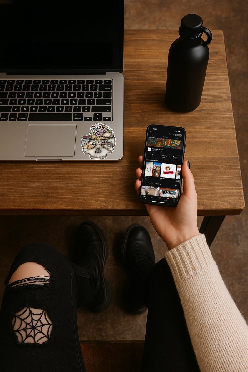 May include: A laptop, a black water bottle, and a smartphone rest on a wooden table. A sticker of a skull with floral accents is on the laptop. The smartphone displays an e-commerce app. The person is wearing black jeans and boots.