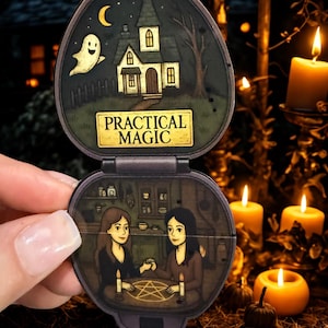 May include: A vintage-style compact with a spooky theme. The top half features a haunted house, ghost, and the words "PRACTICAL MAGIC." The bottom half shows two women at a table with candles and a pentagram. The compact is held open.