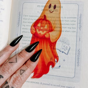 May include: A translucent orange ghost-shaped sticker holding a carved pumpkin. The sticker is on a page with text that reads "MY FAVORITE MAGICAL JOURNALING SHOPS". The ghost has a smiling face. The image is on a book page.