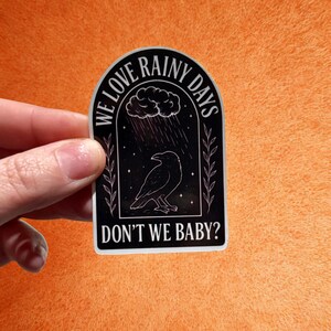 Op de afbeelding: Zwart-witte sticker met de tekst "WE LOVE RAINY DAYS DON'T WE BABY?" met een raaf onder een wolk met regen. De sticker heeft een zwarte achtergrond en een witte rand, gehouden tegen een oranje achtergrond.