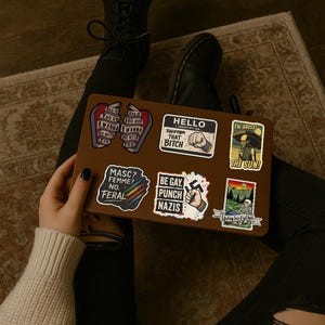 May include: A brown laptop covered in stickers with various designs and text. The stickers include phrases like "Hello, Identify as that Bitch", "Be Gay, Punch Nazis", and "Masc? Femme? No. Feral."