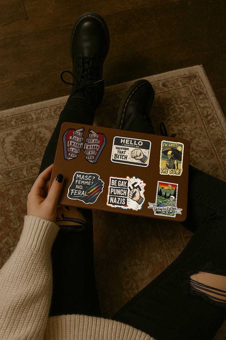May include: A brown laptop covered in stickers with various designs and text. The stickers include phrases like "Hello, Identify as that Bitch", "Be Gay, Punch Nazis", and "Masc? Femme? No. Feral."