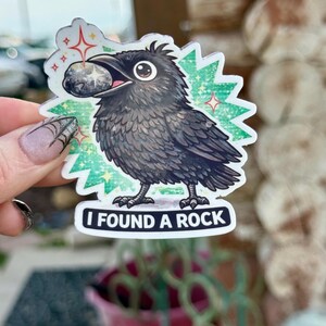 Op de afbeelding: Een sticker met een cartoon kraai die een steen in zijn snavel houdt. De kraai is zwart met gedetailleerde veertjes, tegen een groen-witte achtergrond met de tekst "I FOUND A ROCK".