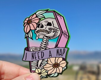 Pode incluir: Um adesivo com um esqueleto em um caixão rosa com borda verde, rodeado por flores. A faixa diz "I NEED A NAP". O fundo é uma imagem desfocada de um céu azul e uma paisagem verde.