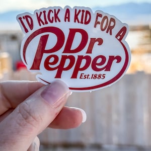 May include: A white sticker with red text that reads "I'd kick a kid for a Dr Pepper Est. 1885." The sticker is held up by a hand against a blurred outdoor background.