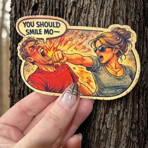 Könnte beinhalten: Ein Aufkleber mit einer Illustration im Retro-Comic-Stil. Das Bild zeigt eine Frau, die einen Mann schlägt. Die Sprechblase enthält den Text "YOU SHOULD SMILE MO-". Der Aufkleber wird in einer Hand vor einem Baumstamm gehalten.