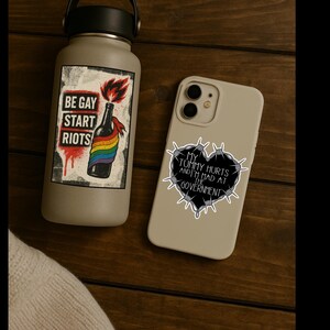 May include: A gray water bottle and a phone case with stickers. The water bottle has a sticker that says "BE GAY START RIOTS" with a rainbow and a bottle. The phone case has a sticker that says "MY TUMMY HURTS AND I'M MAD AT THE GOVERNMENT".