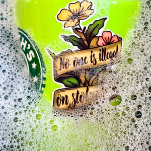 May include: A sticker with a floral design and the text "No one is illegal!" and "on stolen land" on a banner. The sticker features a yellow flower, pink flower, and green leaves. The sticker is on a green surface with bubbles.