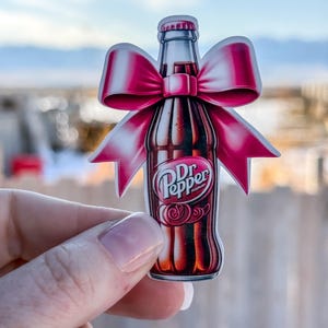 May include: A Dr. Pepper bottle sticker with a pink bow. The sticker features the Dr. Pepper logo in white on a dark brown bottle. The bow is a bright pink color. The background is a blurred outdoor scene.