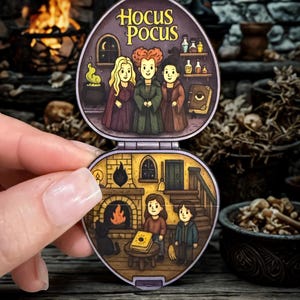 Könnte beinhalten: Ein Miniatur-Klapp-Compact mit Illustrationen aus dem Film "Hocus Pocus". Die obere Hälfte zeigt die Sanderson-Schwestern, während die untere Hälfte eine Szene mit einem Kamin und Kindern darstellt. Der Compact wird vor einem verschwommenen Hintergrund gehalten.