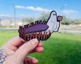 Pode incluir: Um autocolante com tema de Halloween com um fantasma de desenho animado num caixão. O fantasma branco com olhos pretos segura uma placa que diz "BRB". O caixão é roxo, rodeado por uma cerca de madeira castanha. Fundo: relva verde e céu azul.