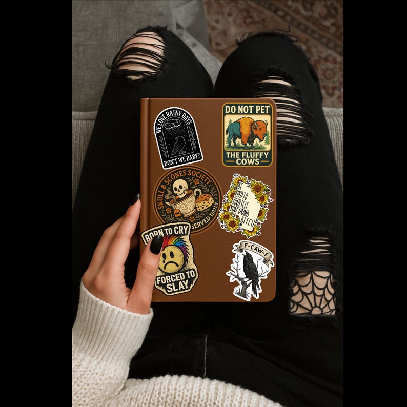 May include: A brown journal covered in stickers, including phrases like "We Love Rainy Days," "Do Not Pet," and "Forced to Slay." The journal is held by a person wearing a white sweater and black ripped jeans.
