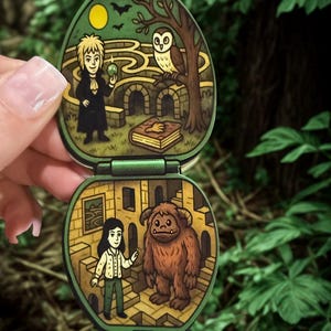 Op de afbeelding: Een groen, ovaal speelgoed met twee scharnierende secties, elk met een gedetailleerde, geïllustreerde scène. Het bovenste gedeelte toont een doolhof met een figuur, een uil en een boek. Het onderste gedeelte toont een meisje en een harig wezen.