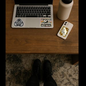 May include: A laptop with stickers, a beige water bottle, and a phone case with a sunflower design are on a wooden surface. The stickers read "WE ARE THE WEIRDOS MISTER" and "OUT THE CLOSET INTO THE PIT". The phone case says "SHOULD'VE KNOWN I'M A BITCH".
