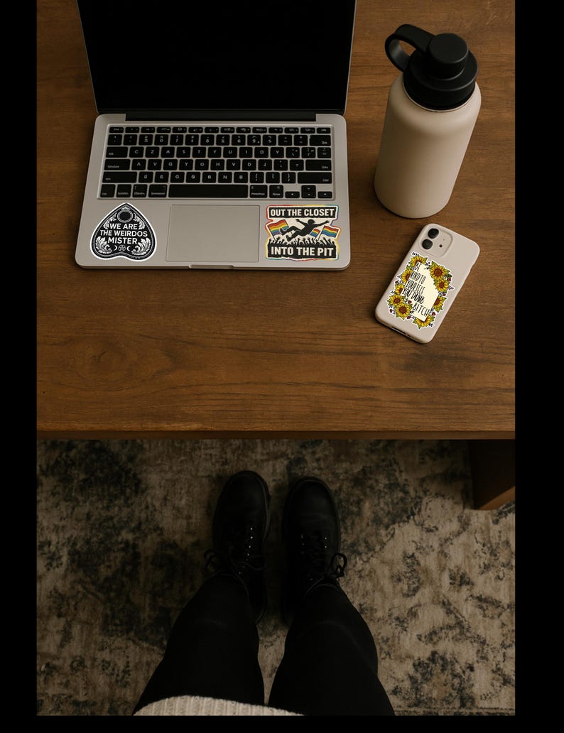 May include: A laptop with stickers, a beige water bottle, and a phone case with a sunflower design are on a wooden surface. The stickers read "WE ARE THE WEIRDOS MISTER" and "OUT THE CLOSET INTO THE PIT". The phone case says "SHOULD'VE KNOWN I'M A BITCH".