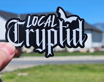 Pode incluir: Um autocolante preto com as palavras "LOCAL Cryptid" em branco, com letras góticas estilizadas. O autocolante é segurado contra um céu azul e relva verde.
