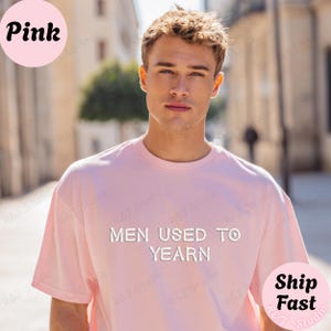 May include: A light pink short-sleeved t-shirt featuring the white text "MEN USED TO YEARN" printed across the chest. The garment has a classic crew neckline and a relaxed fit, providing a simple and straightforward design for casual wear.