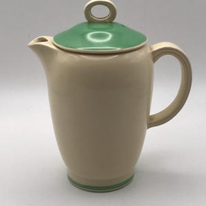 May include: Cream-coloured ceramic jug with a green lid and handle. The jug has a rounded body, a spout, and a curved handle. The lid has a small loop handle. A green stripe accents the base. A classic kitchenware item.