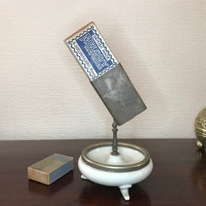 May include: A white ceramic ashtray with a silver rim and a metal stand. A vintage matchbox with the text "British Made Melody Matches H. Sheldon & Sons, Evesham" is attached to the stand.