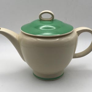 May include: Cream ceramic teapot with a green lid and handle. The teapot has a curved spout and a rounded body. A green stripe accents the base. The lid has a loop handle.
