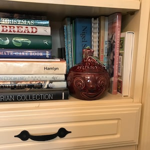 May include: A collection of cookbooks on a shelf, including titles such as "Bread" and "Vegetarian Cooking." A maroon ceramic beetroot-shaped container with a face is placed among the books. The shelf is part of a cream-coloured cabinet.