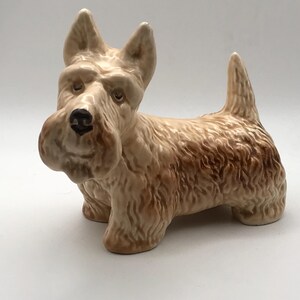 May include: A ceramic ornament of a Scottish Terrier dog. The dog is tan with darker brown shading and a black nose. The figurine has upright ears and a short, upright tail. The dog is standing on all four legs.