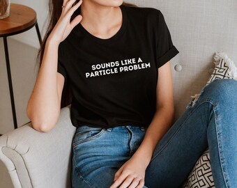 May include: A woman wearing a black t-shirt with the text "Sounds like a particle problem" in white lettering. She is sitting in a chair wearing blue jeans with a distressed knee.
