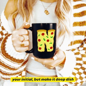 May include: Black coffee mug with the letter "N" designed as a pizza slice, with red and green toppings. The mug has the text "your initial, but make it deep dish" in yellow. The mug is held by a person.