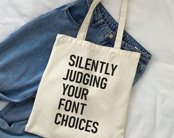 Könnte beinhalten: Eine cremefarbene Tragetasche mit schwarzem Text, der "SILENTLY JUDGING YOUR FONT CHOICES" lautet. Die Tasche hat zwei Griffe und liegt auf einer Jeans. Der Hintergrund ist ein weißes Laken.