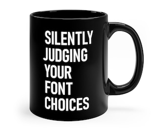 Könnte beinhalten: Schwarze Keramik-Tasse mit weißem Text: "SILENTLY JUDGING YOUR FONT CHOICES". Die Tasse hat einen gebogenen Henkel und eine glänzende Oberfläche. Geeignet für Heißgetränke.