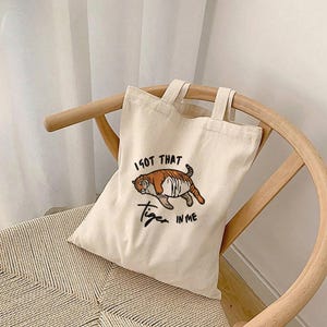 May include: A cream-colored tote bag with a tiger illustration and the text "I GOT THAT Tiger IN ME". The tiger is brown and orange. The bag has two handles and is sitting on a wooden chair.