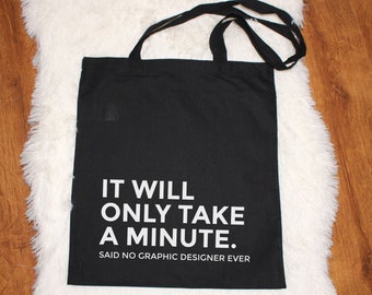 Könnte beinhalten: Schwarze Tragetasche mit schwarzen Henkeln. Die Tasche hat weißen Text, der lautet: "IT WILL ONLY TAKE A MINUTE. SAID NO GRAPHIC DESIGNER EVER."