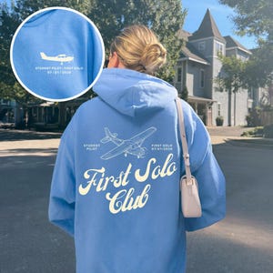 May include: A light blue hooded sweatshirt featuring a white graphic of a small aeroplane on the back, accompanied by the text "STUDENT PILOT" and "FIRST SOLO 07/01/2026" above the words "First Solo Club" in a large, cursive white font.