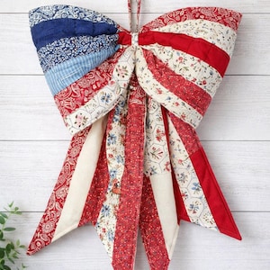 May include: A quilted fabric bow decoration featuring a patchwork design of red, white, and blue patterns. The bow includes floral, paisley, and striped fabric segments with long, pointed tails hanging downward, suspended by a tan fabric loop.