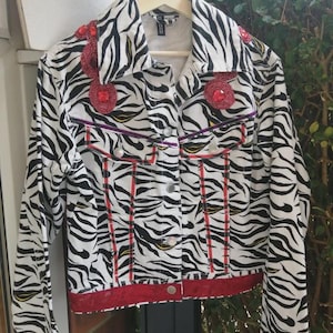 May include: A long-sleeve denim jacket featuring a black and white zebra print pattern. The jacket is embellished with circular red beaded patches on the chest and shoulders, and includes red fabric trim along the bottom hem and sleeve cuffs.